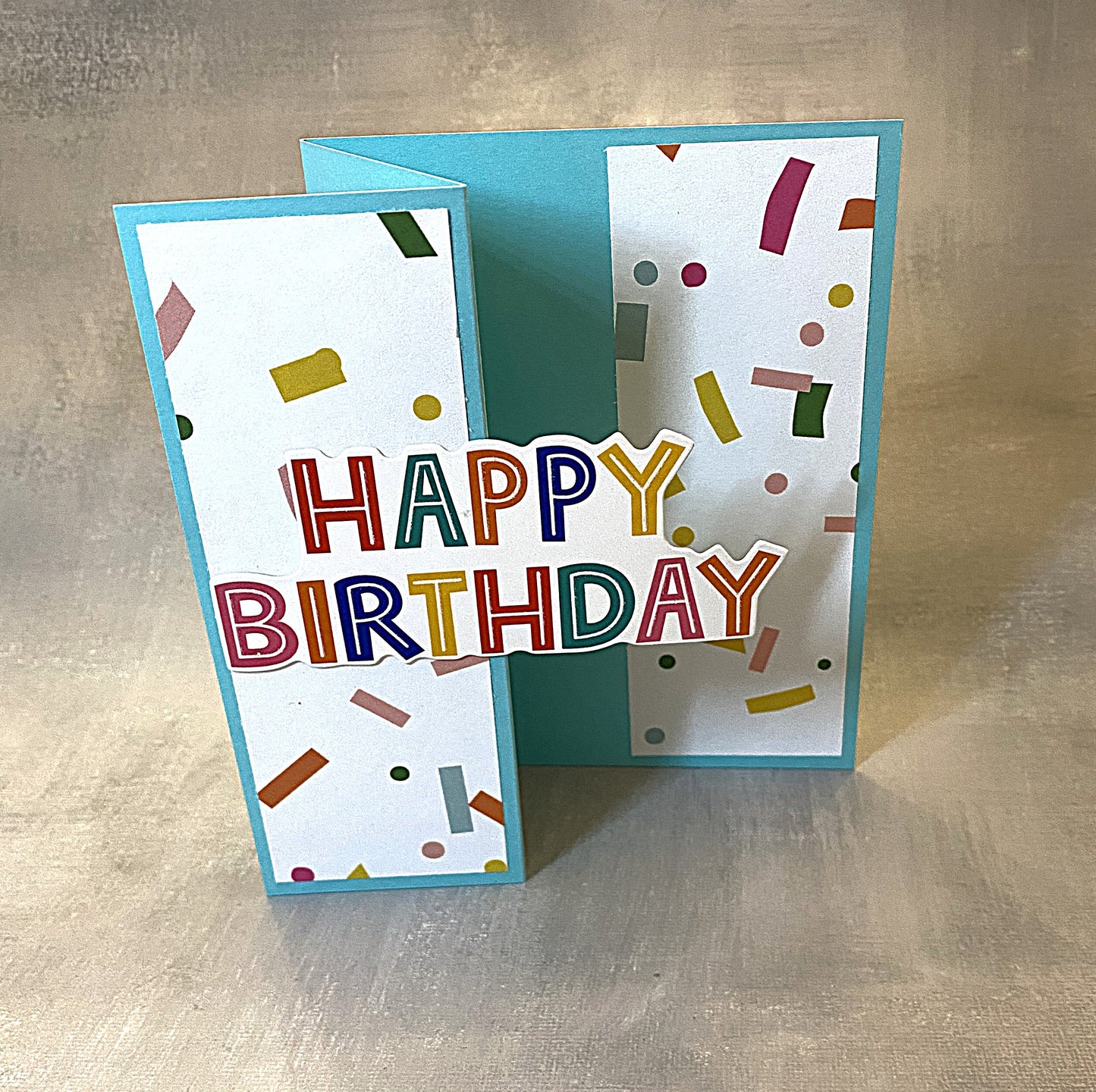 Aqua blue bi-fold birthday card featuring colorful confetti and “Happy Birthday” on the front, blank inside.