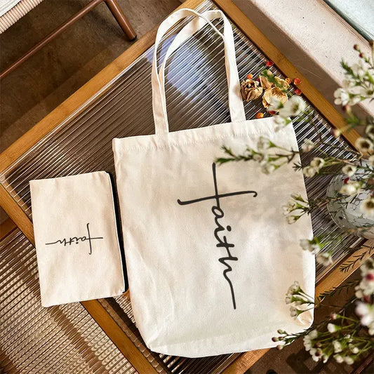 Cream canvas tote bag with the word "Faith" printed vertically down the front, accompanied by a matching small zipper pouch (9" x 6") with the same design. Tote measures 15" tall x 13.5" wide