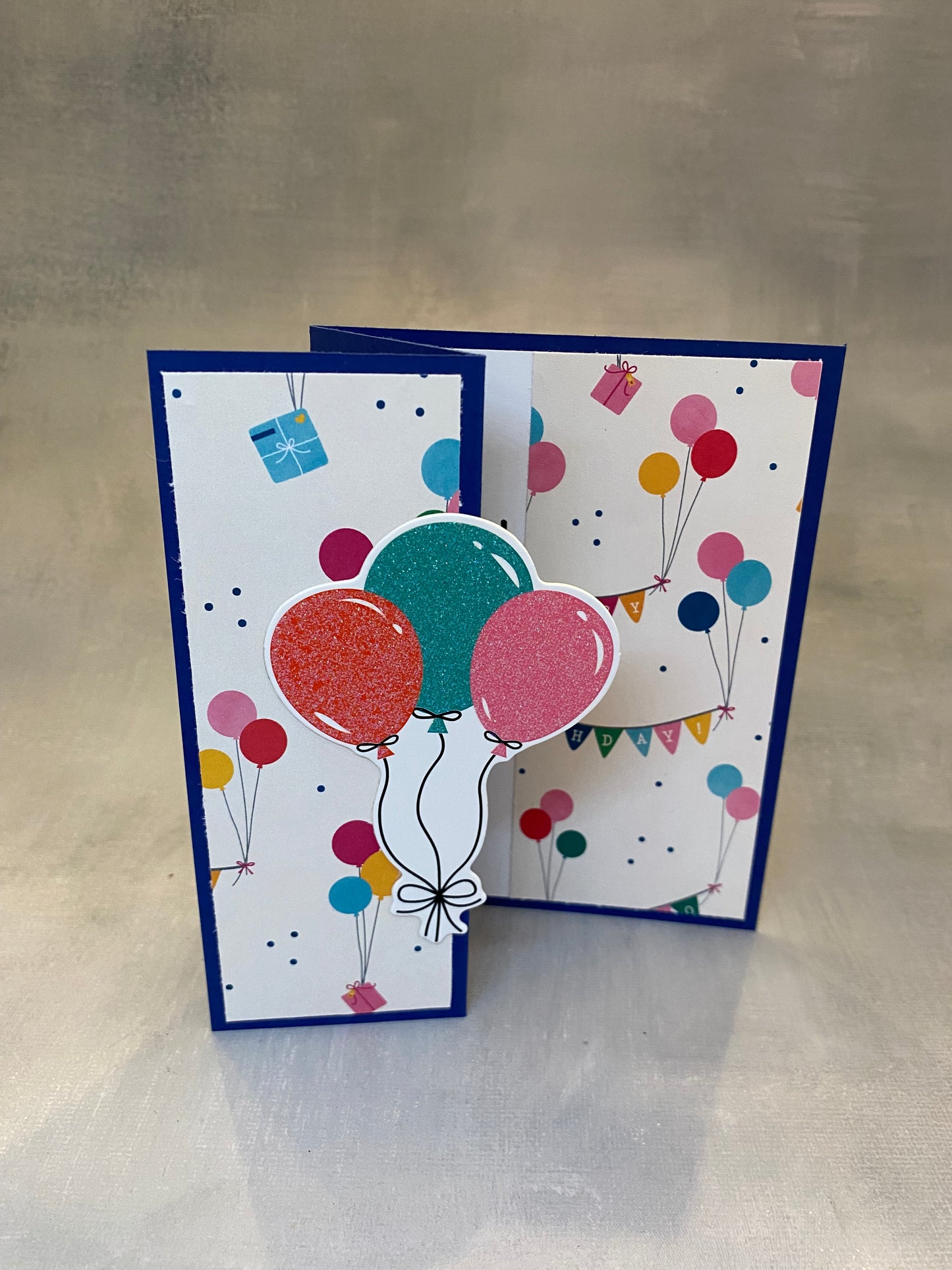 "Dark blue birthday card featuring a colorful party scene and balloon bouquet, displayed with a white envelope."