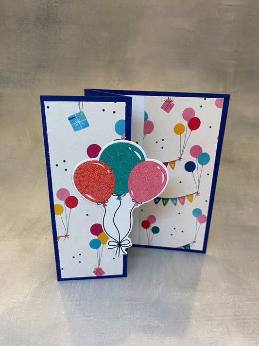 "Dark blue birthday card featuring a colorful party scene and balloon bouquet, displayed with a white envelope."