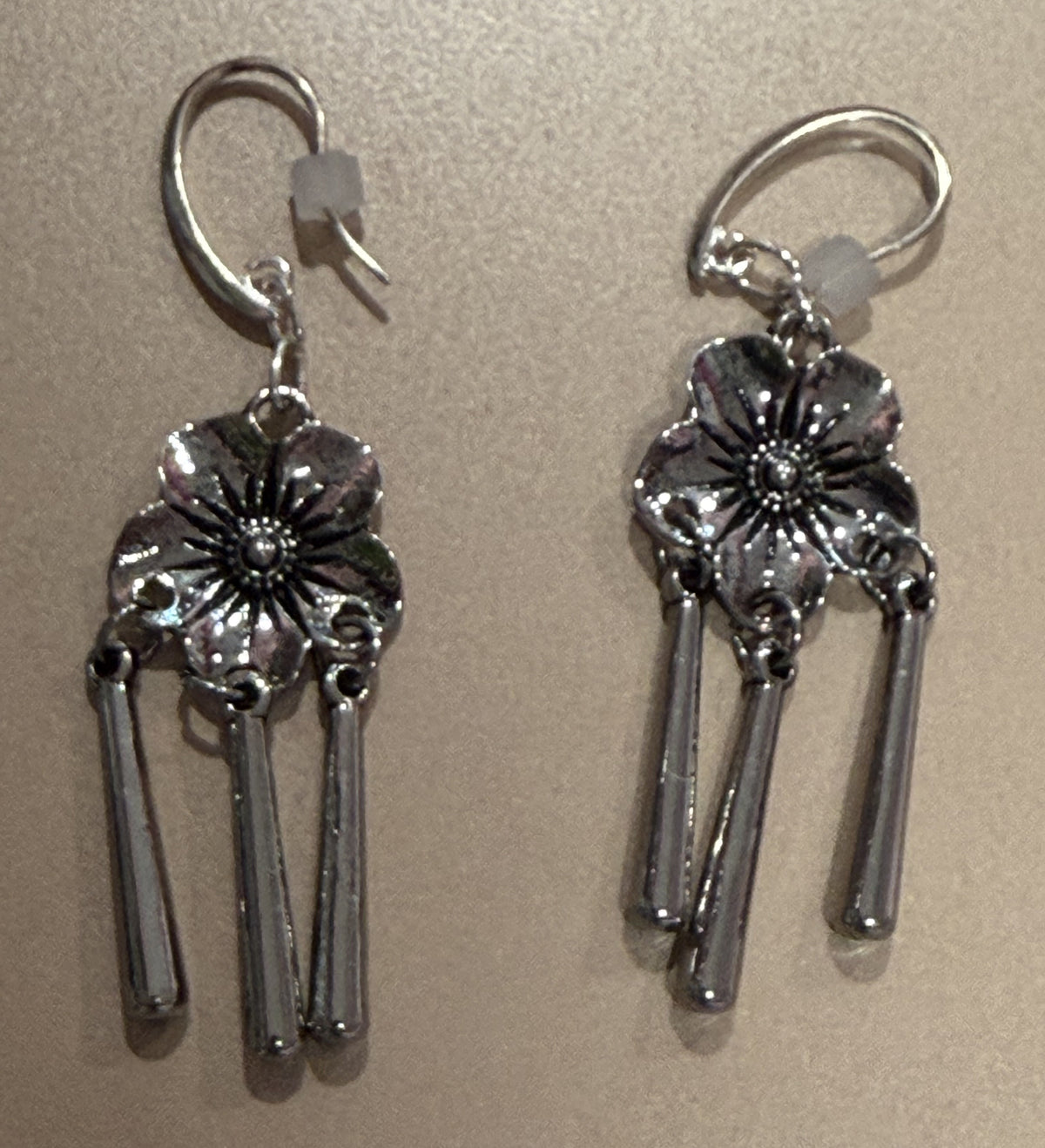A pair of silver-toned dangle earrings with flower charms and three hanging bars, shown on a plain beige background.