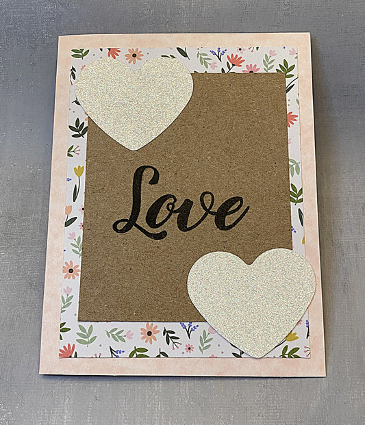 Pink greeting card with hand-stamped 'Love' on the front, decorated with pink flowers and glitter heart accents, shown with a white envelope."