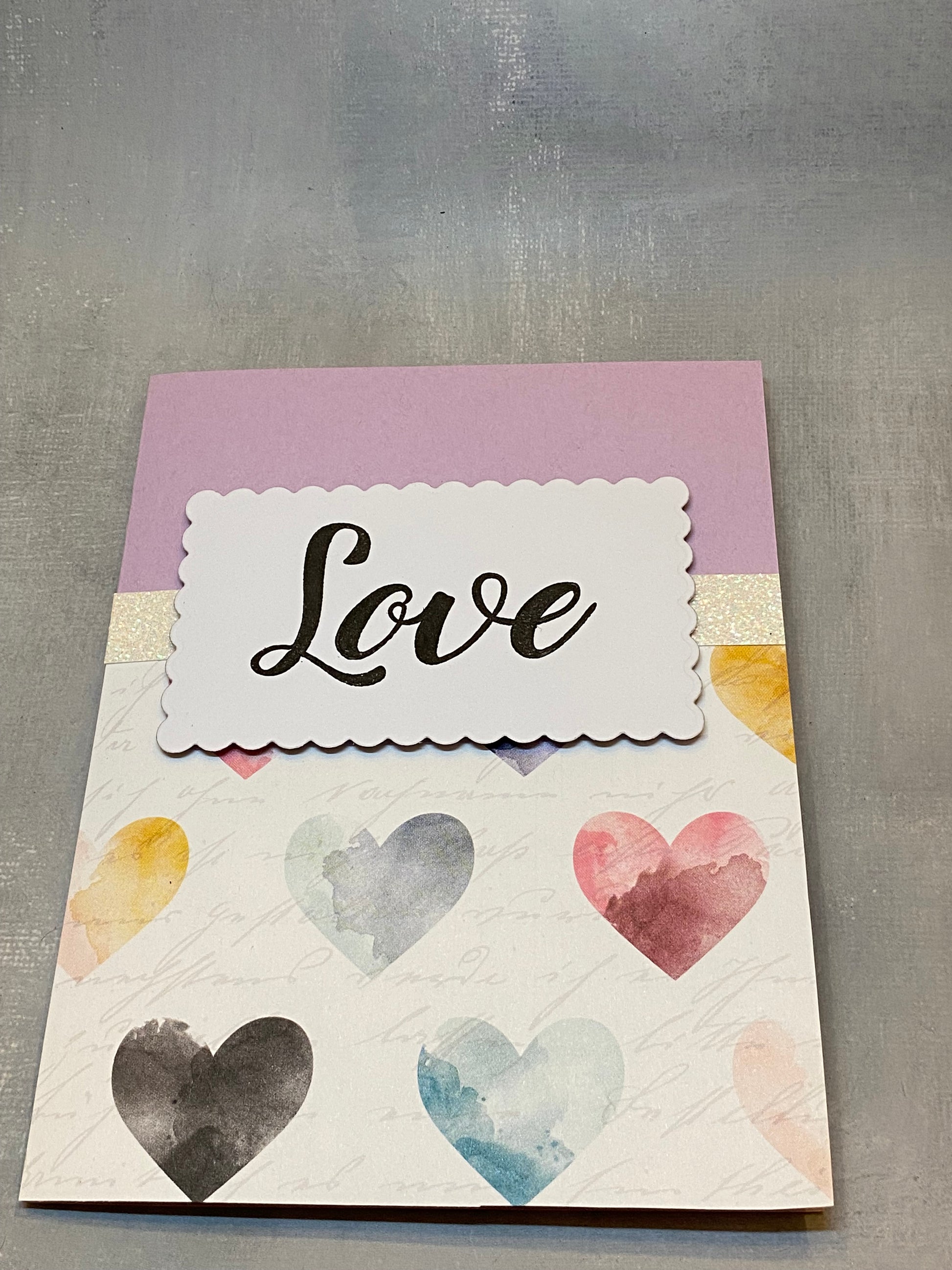 "Purple anniversary card with hand-stamped 'Love' on the front, featuring muted watercolor hearts and glitter accents, displayed with a white envelope."