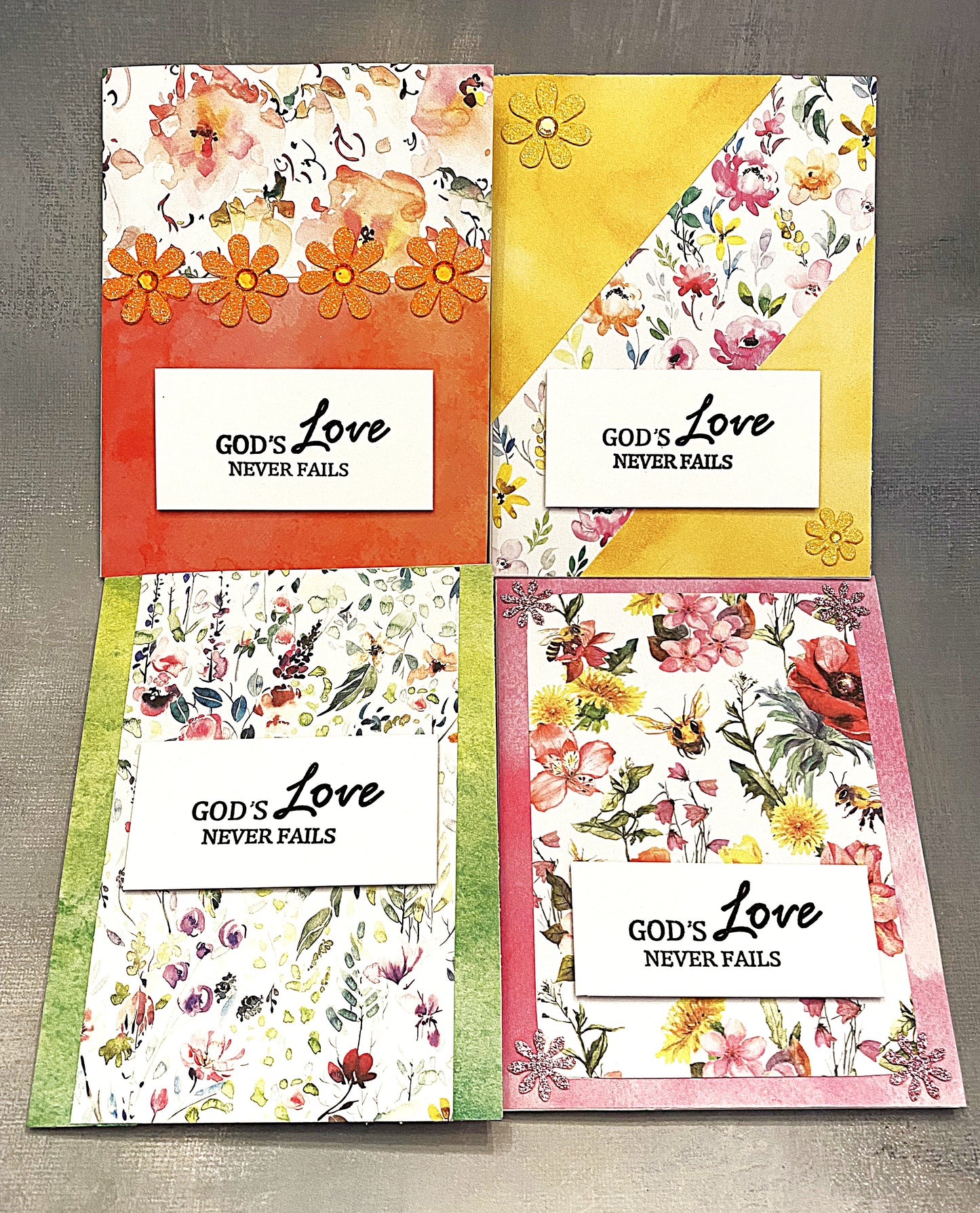 Set of four 4¼" x 5½" floral note cards with a watercolor effect, hand-stamped with “God’s Love Never Fails” on the front, each paired with a white envelope.







