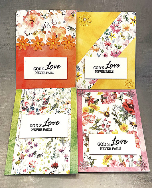 Set of four 4¼" x 5½" floral note cards with a watercolor effect, hand-stamped with “God’s Love Never Fails” on the front, each paired with a white envelope.








