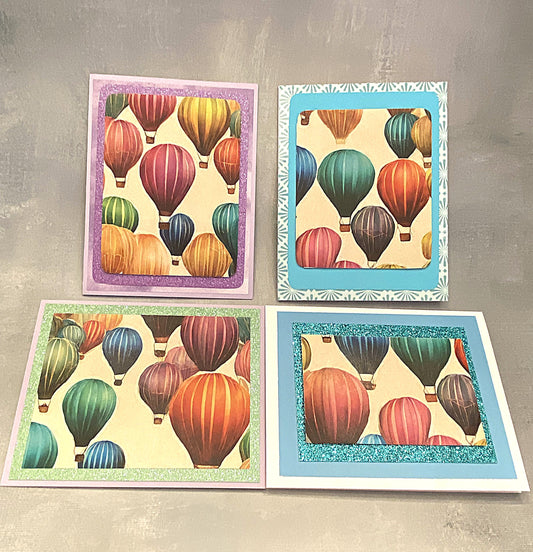 "Set of four 4¼ x 5½ inch note cards featuring colorful hot air balloons with glitter paper accents, each paired with a matching envelope, displayed on a flat surface."