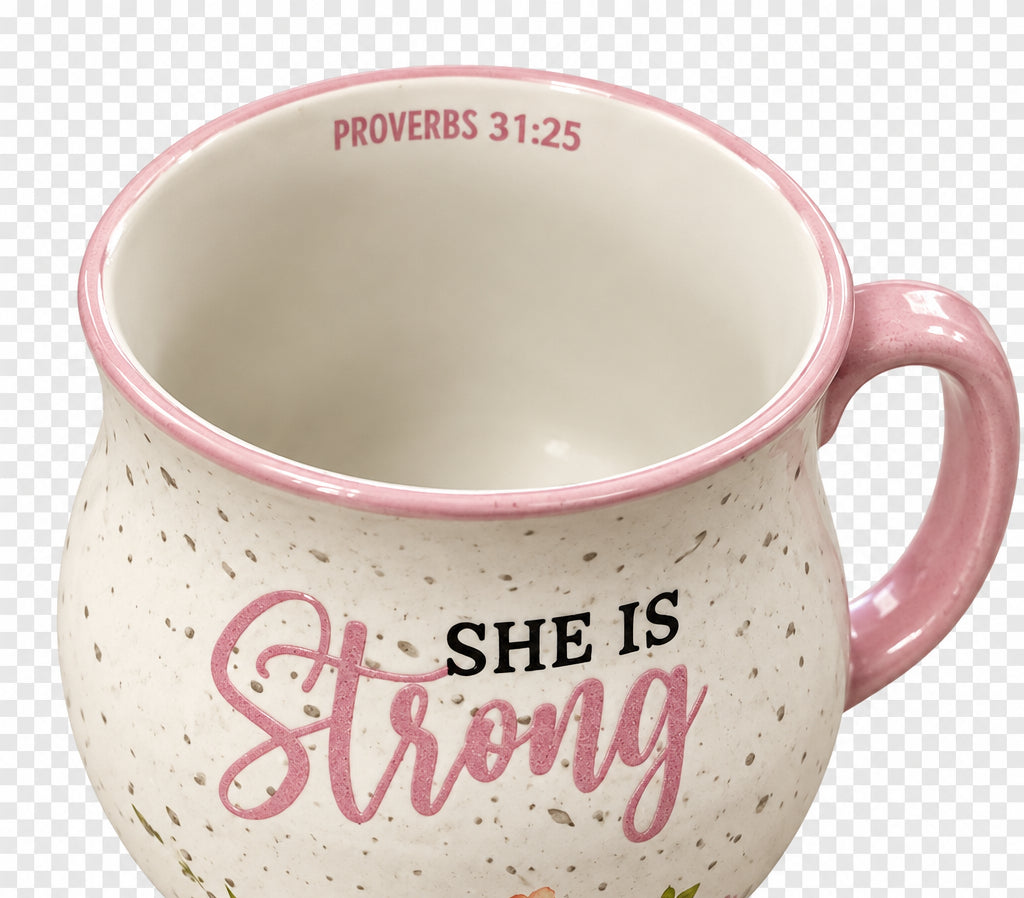 A ceramic mug with a speckled white exterior, featuring the words 'SHE IS Strong' in black and dusty pink lettering, with 'PROVERBS 31:25' printed inside the rim.