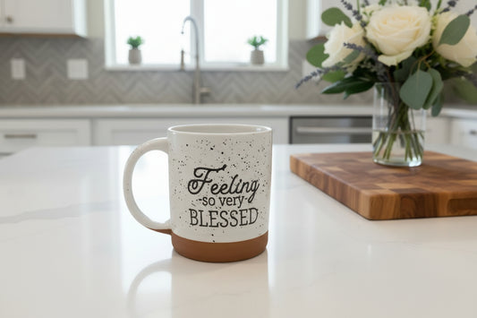 White 12 oz ceramic mug with speckled glaze, “Feeling so very blessed” text, and a natural terra cotta bisque bottom quarter